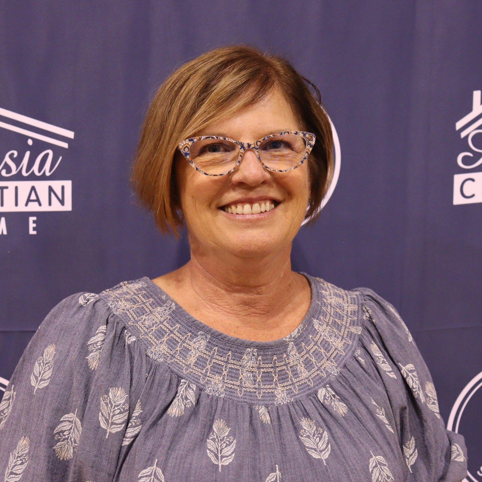 Woman with glasses smiling, wearing a blue patterned blouse in front of a backdrop with logos.