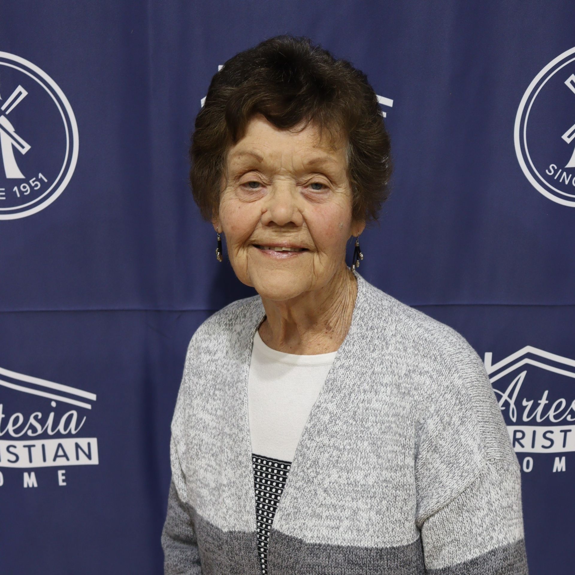 Woman smiling, wearing gray sweater, posing in front of Artesia Christian Home backdrop.