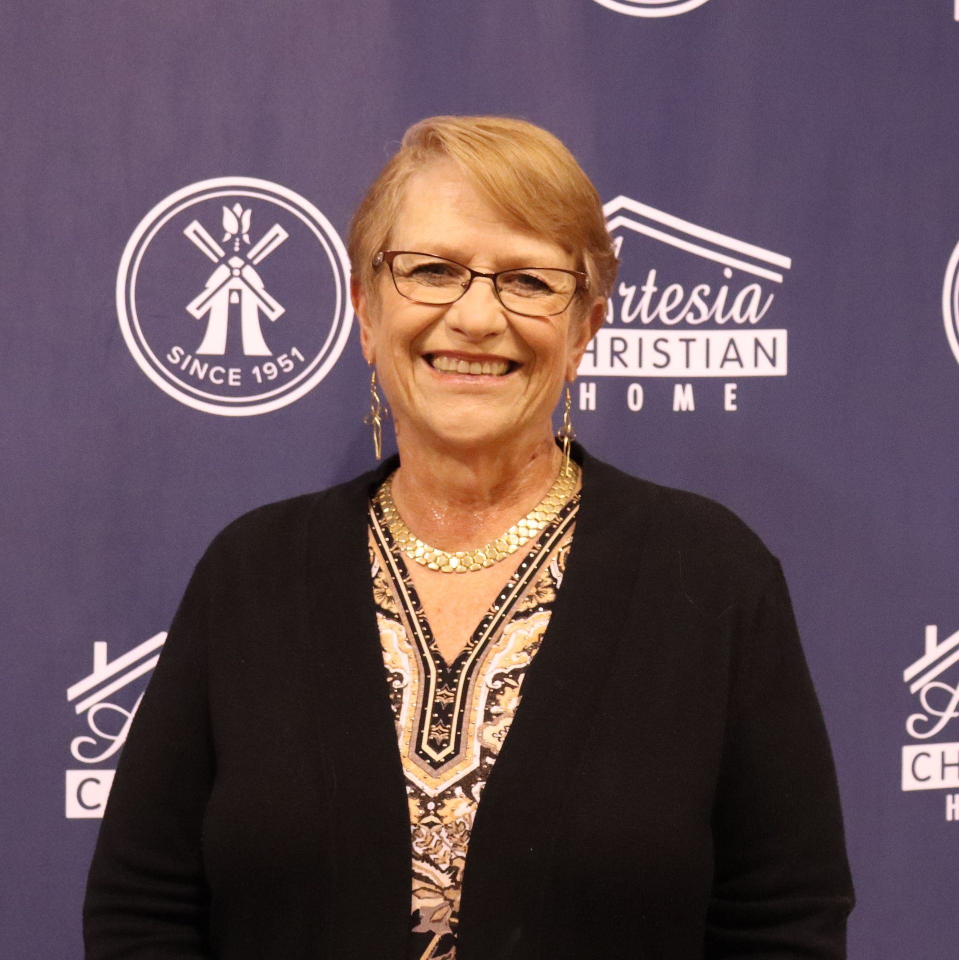 Woman smiling in front of a backdrop with logos; wearing glasses, black cardigan, and a necklace.