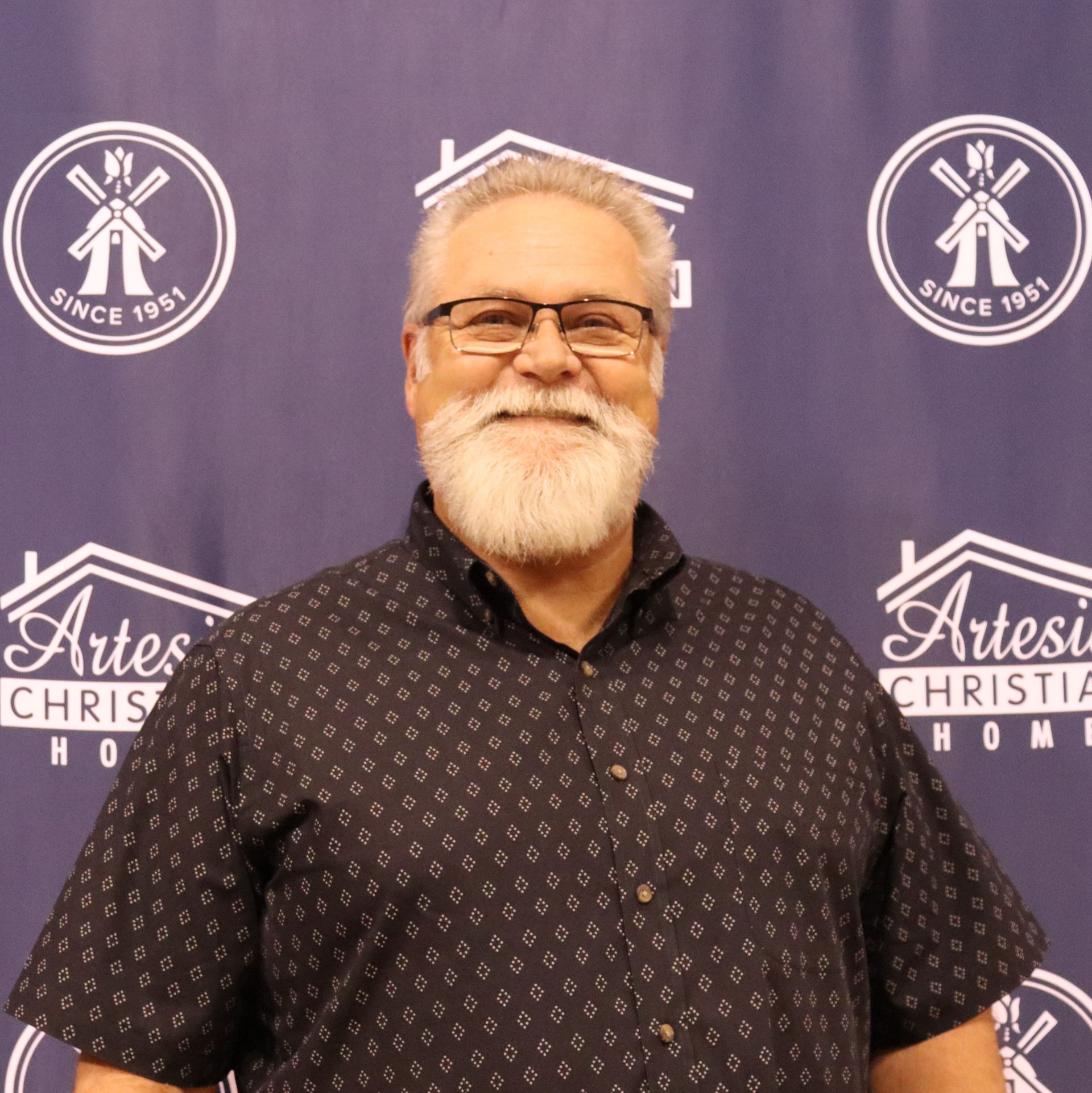 Smiling man with white beard and glasses, standing in front of a backdrop with logos.