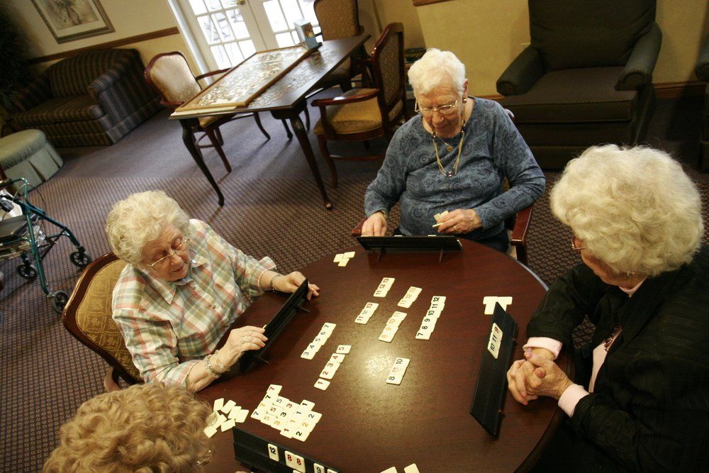 Four elderly women sit at a round table and play dominoes.