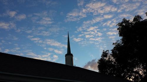 Church steeple silhouetted against a blue sky with scattered white clouds, trees on right.