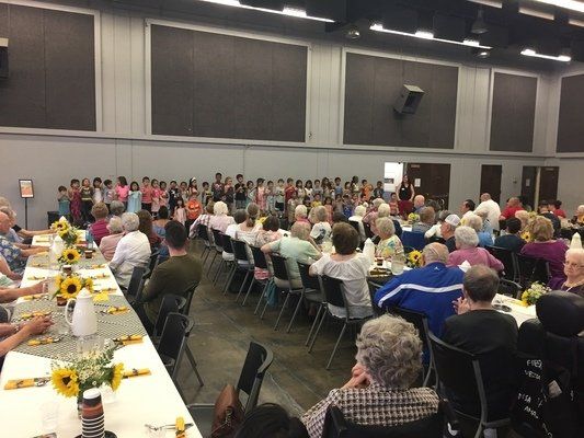 People seated at tables watch a choir performing in a large hall.