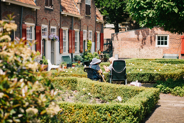Two people sit in chairs in a garden surrounded by hedges; brick buildings in the background.