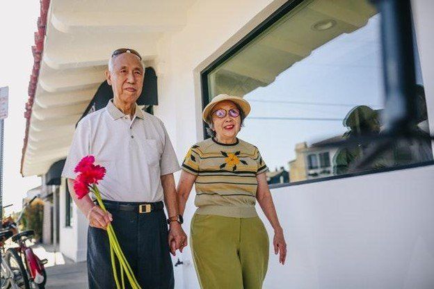Elderly couple, holding hands, walking outside. Man holds red flowers. Sunny day.
