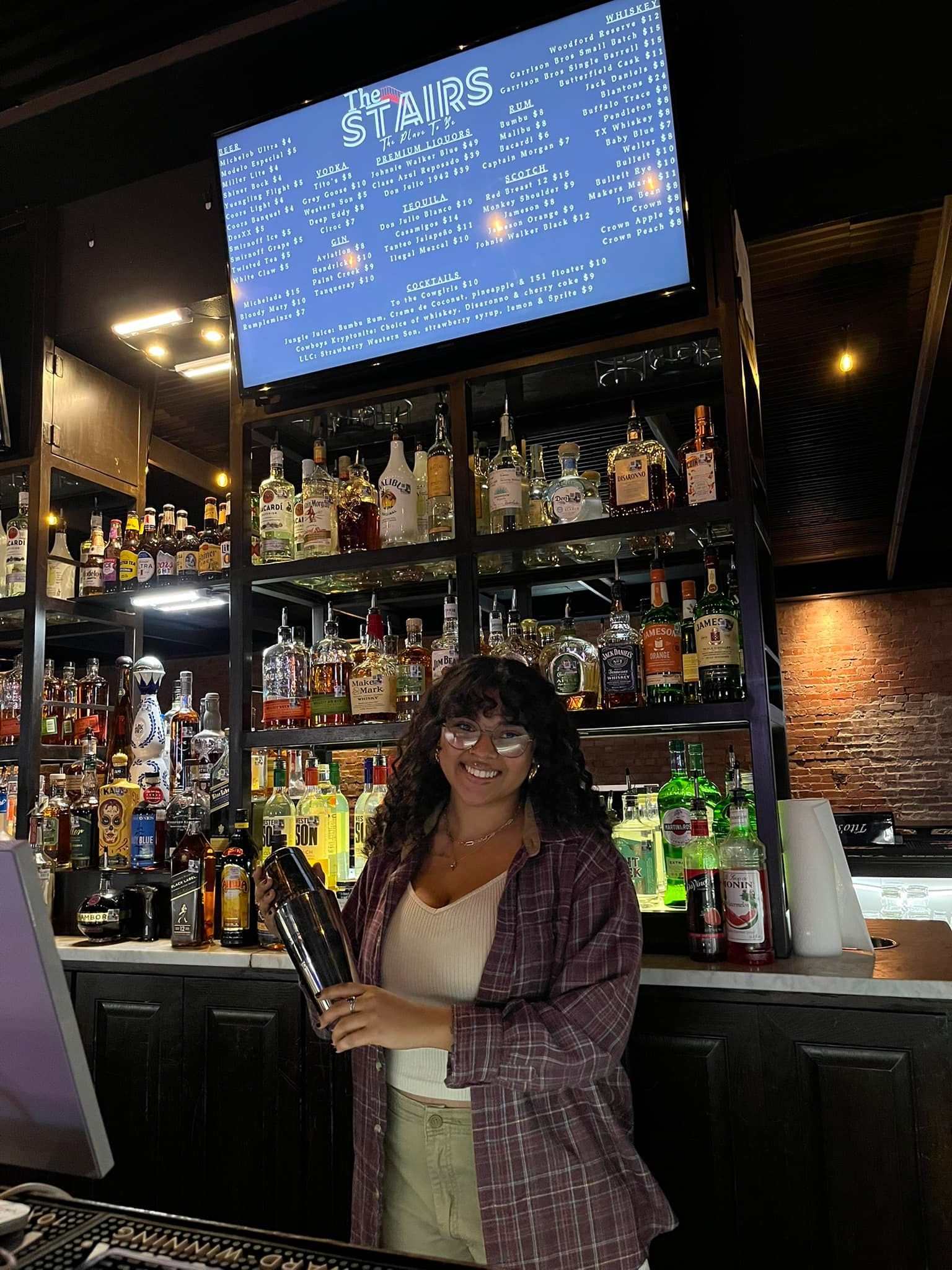 A woman is standing in front of a bar filled with bottles of alcohol.