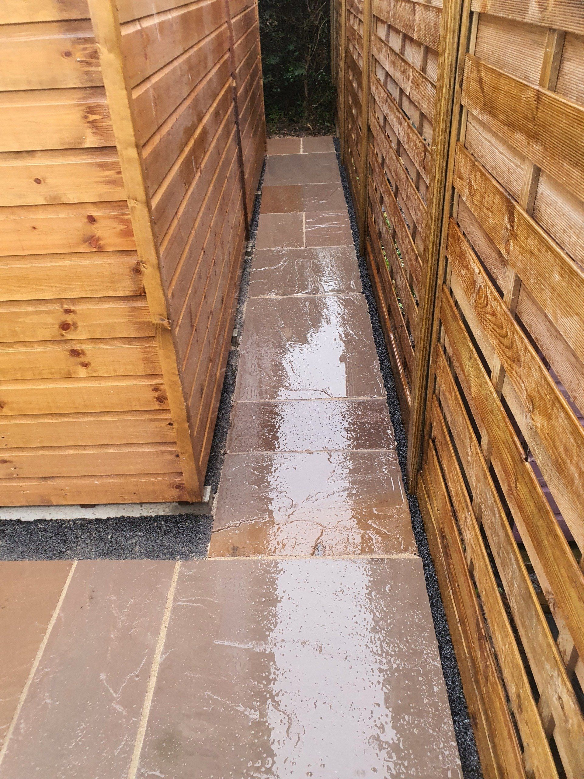 Narrow stone path between a wooden shed and fence. Wet stones, gravel edging, sunlight.