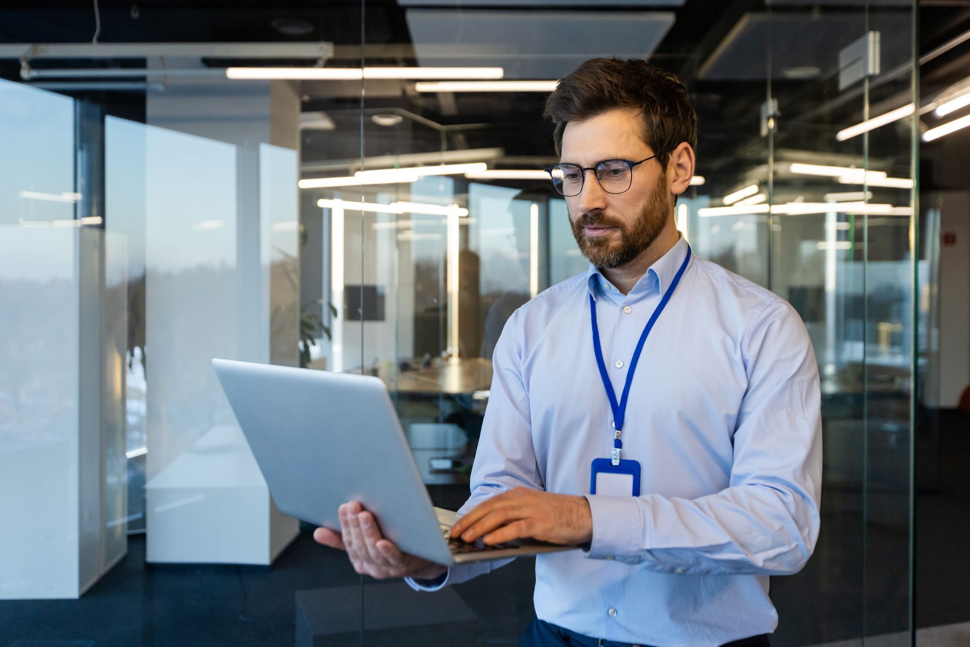 Man in glasses and light blue shirt working on a laptop in a modern office, wearing an ID badge.