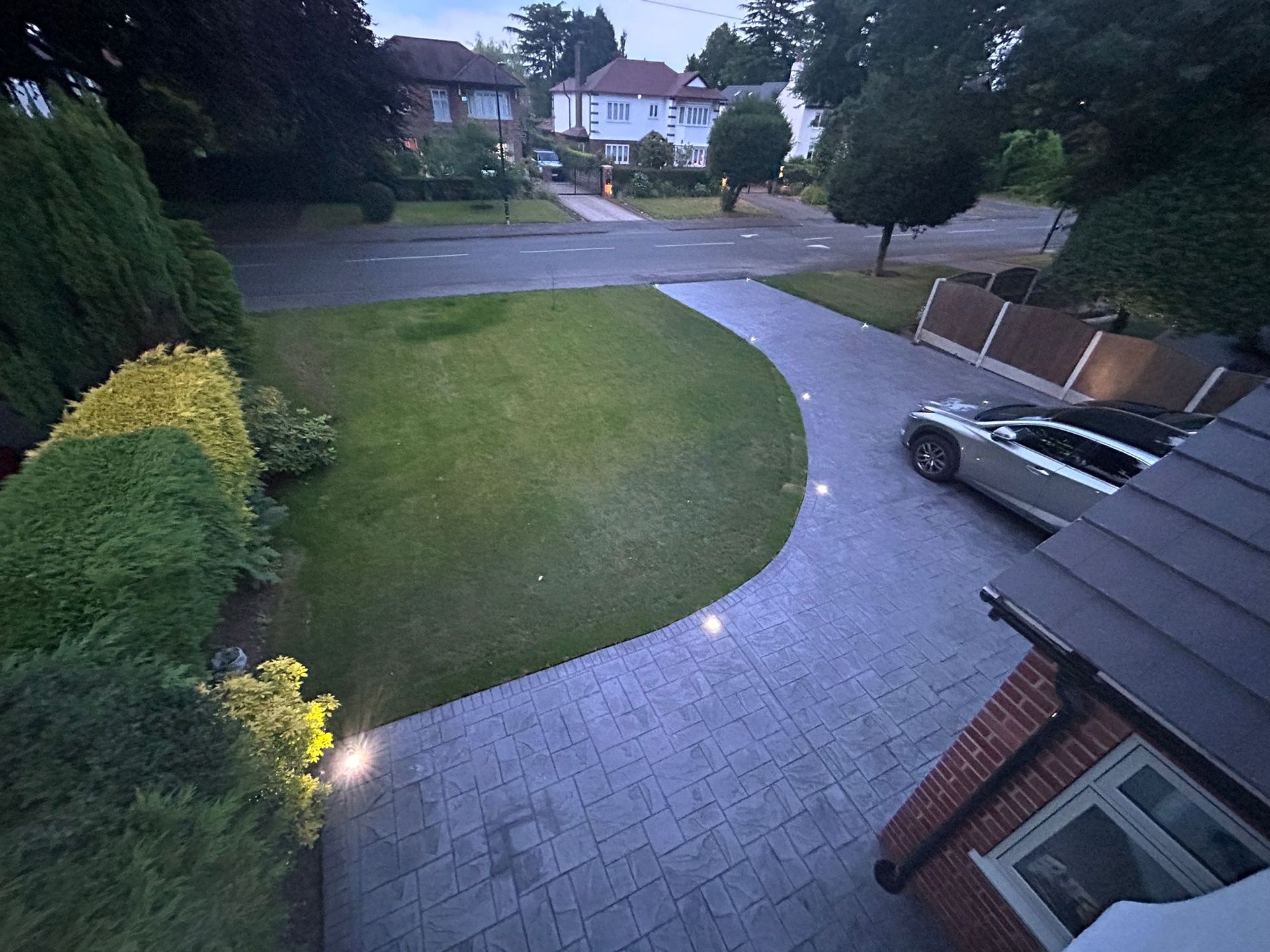 Driveway leading to street, with grass lawn and parked car, lit by ground lights; dusk setting.