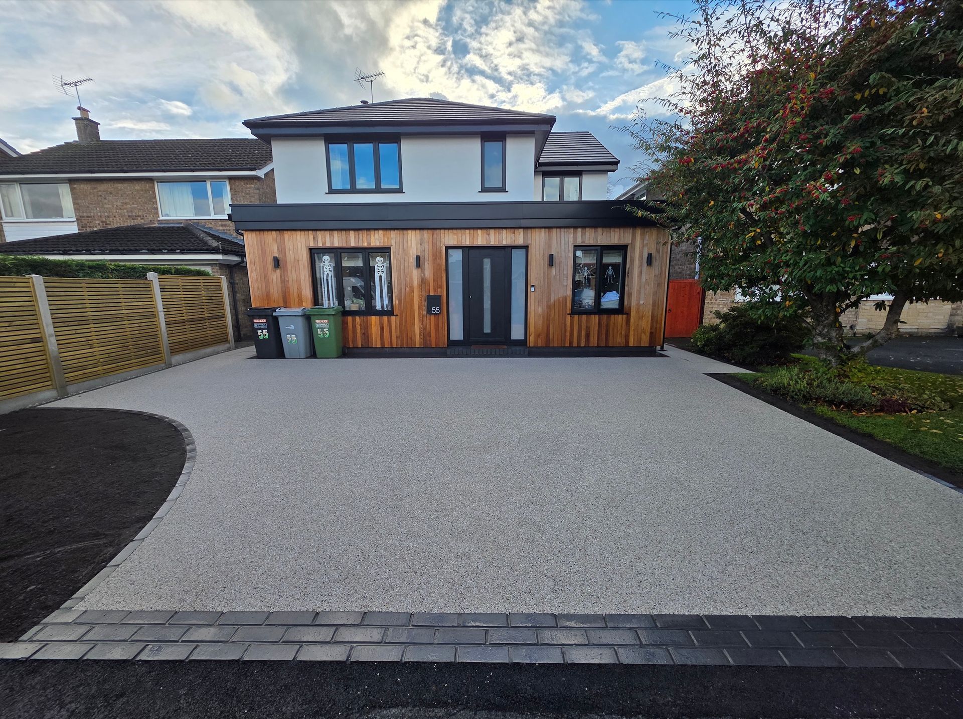 Two-story house with a wooden extension, gravel driveway, and a partially sunny sky.
