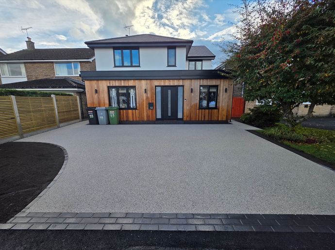 House with gravel driveway and wooden addition; trash bins visible. Blue sky.