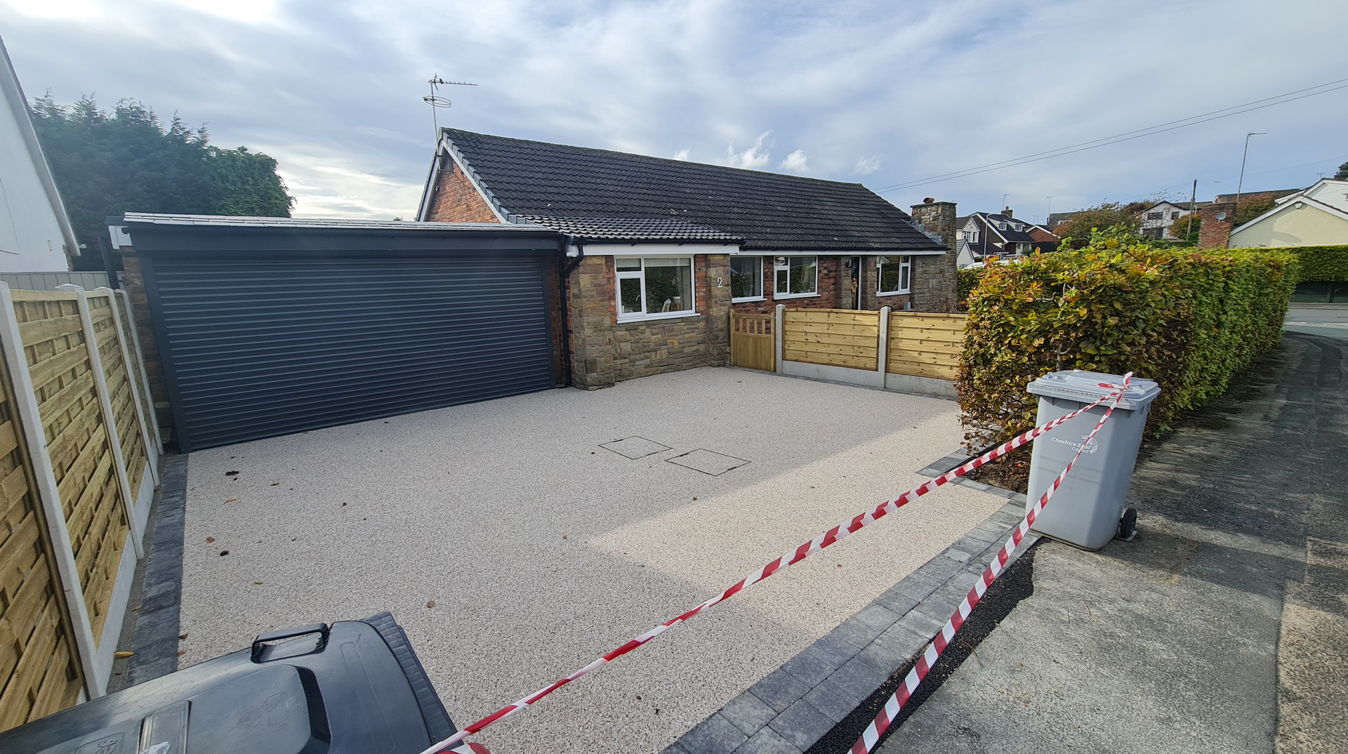 Gravel driveway in front of a brick bungalow with a garage. A wooden fence and green hedges border the property.