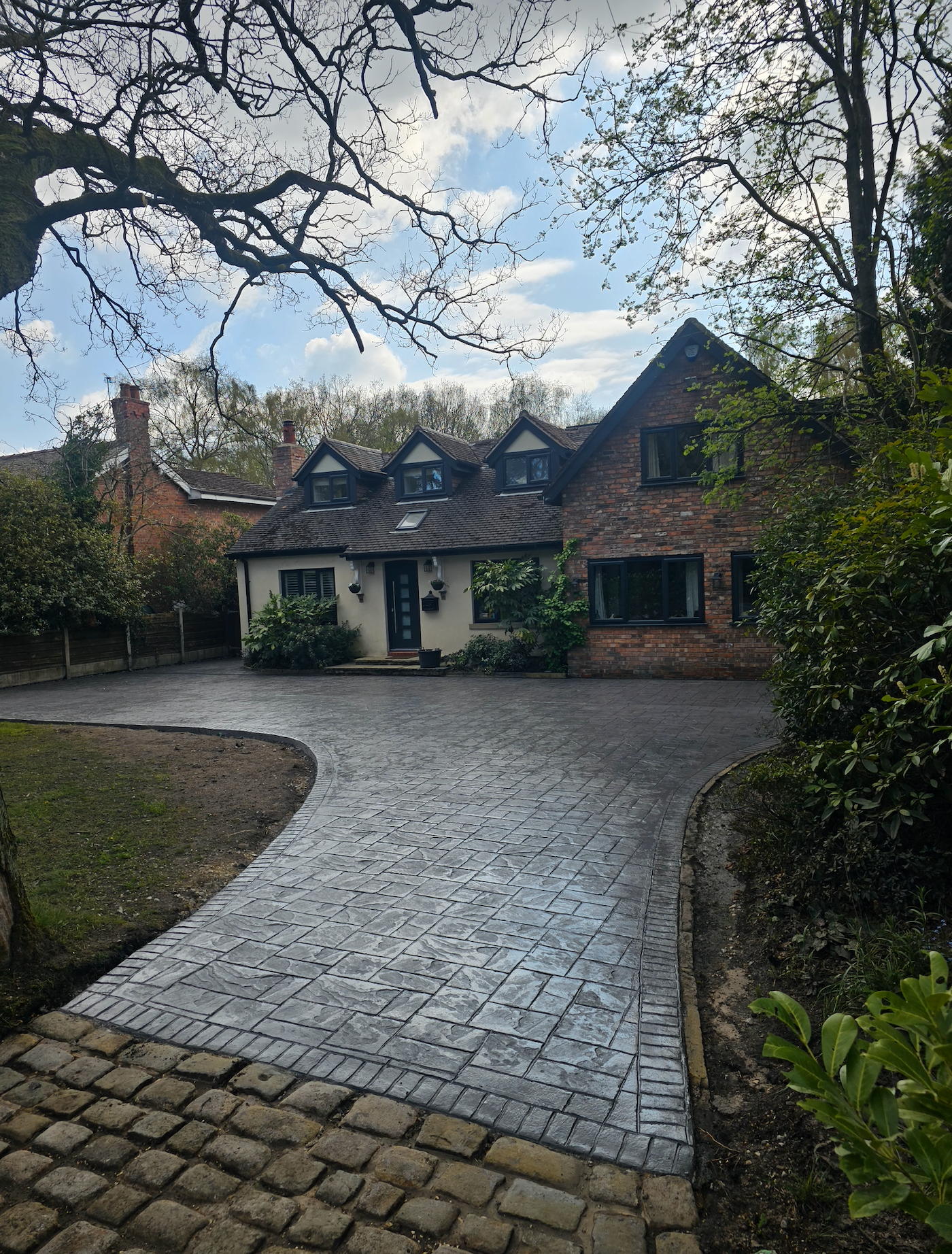 Brick home with cobblestone driveway, surrounded by trees and greenery under a cloudy sky.