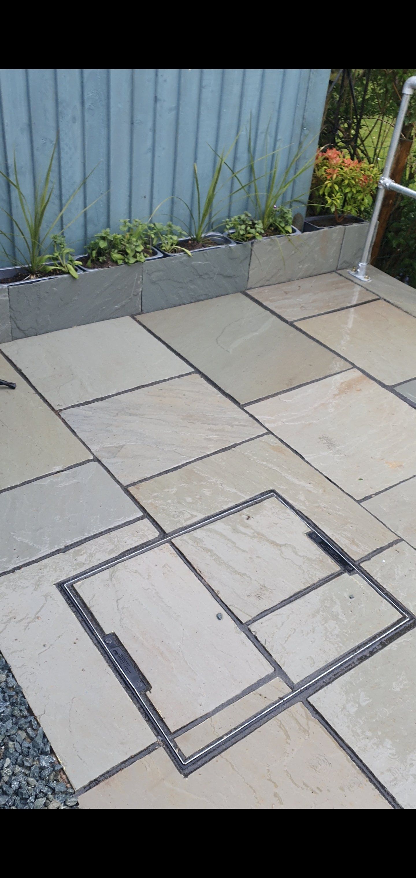 Patio with light-colored paving stones and a raised bed with plants, a metal access panel in the foreground.