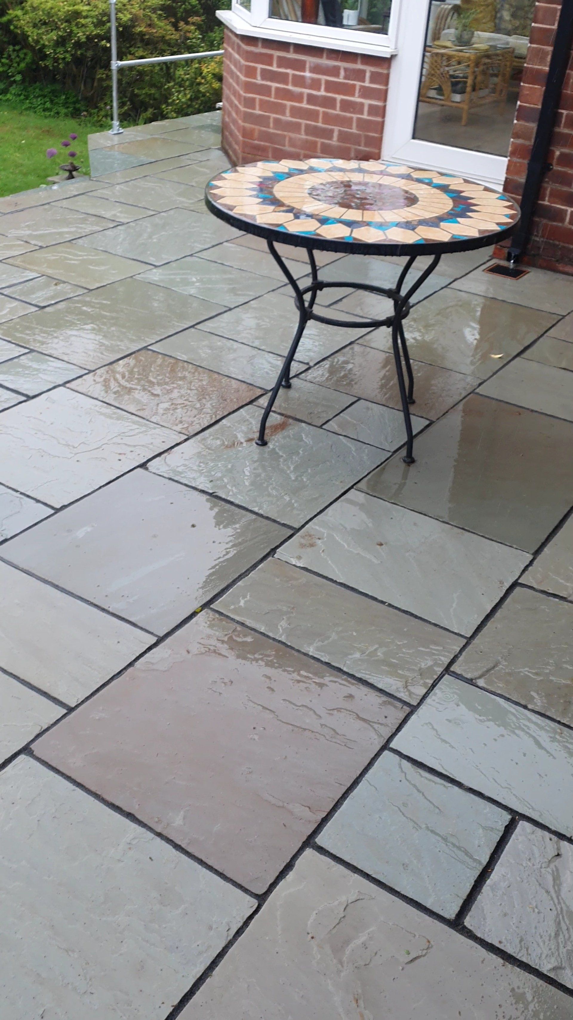 Round mosaic table on a wet paved patio. Brick building background and green grass visible.