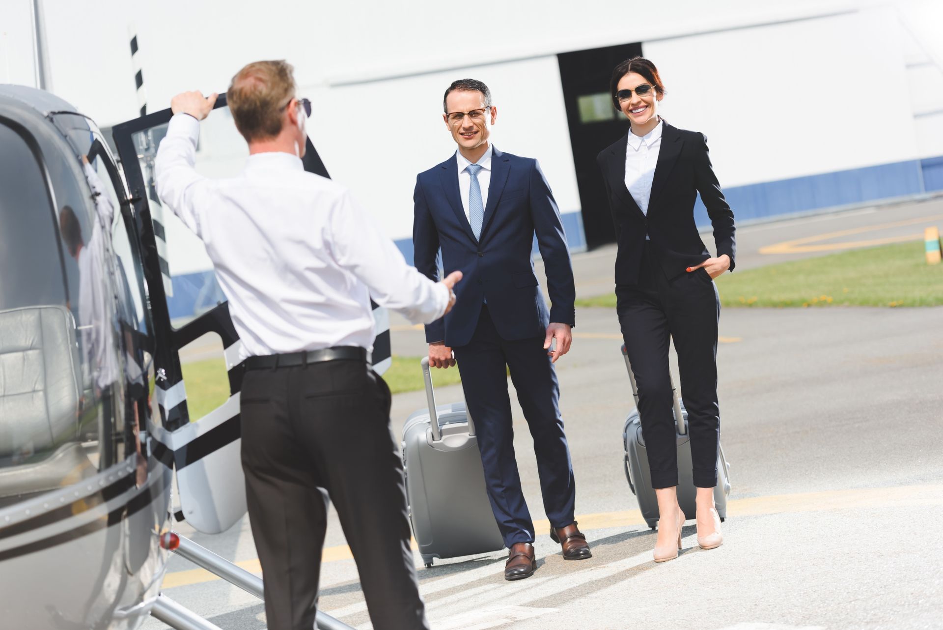 Man in uniform helps two people with luggage near a helicopter on a helipad.
