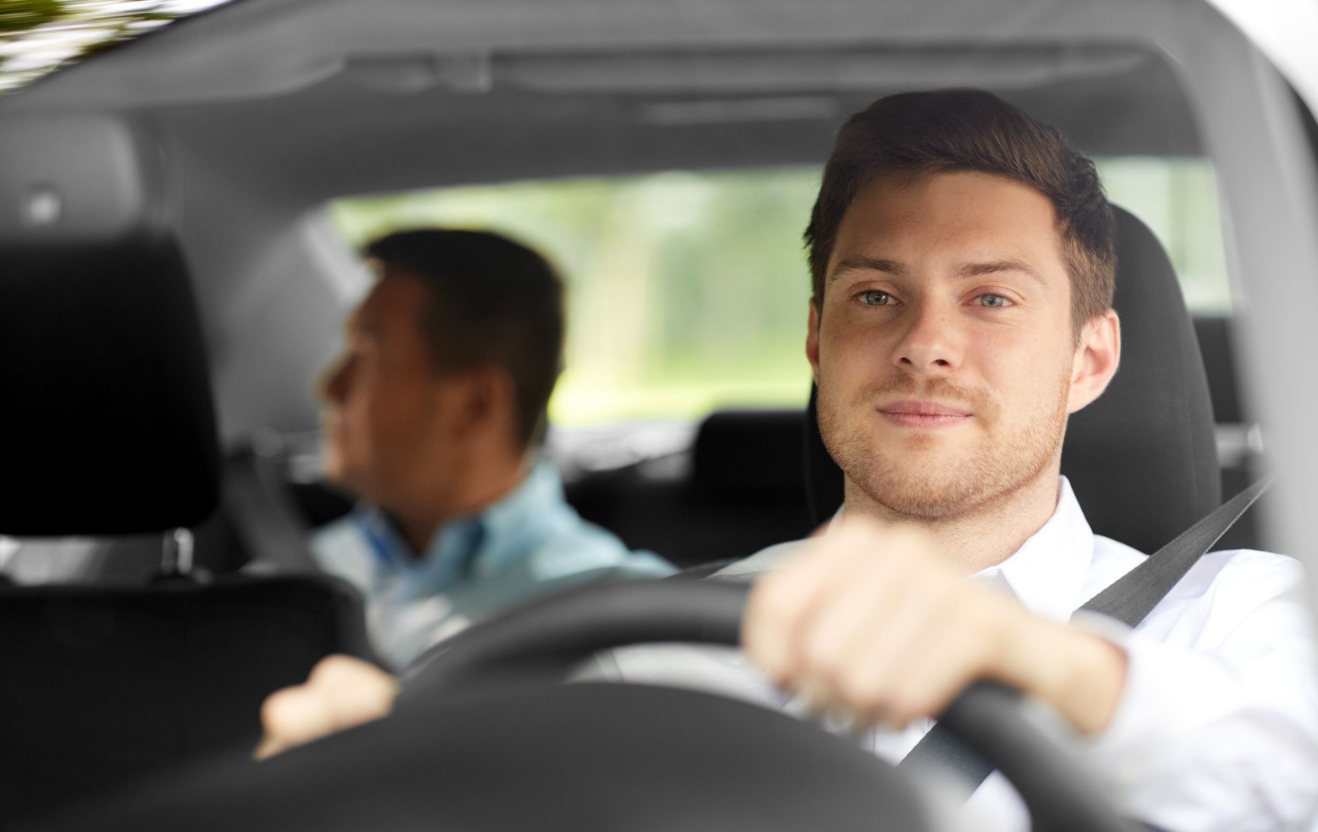 Man driving a car, smiling, with a passenger in the back seat.