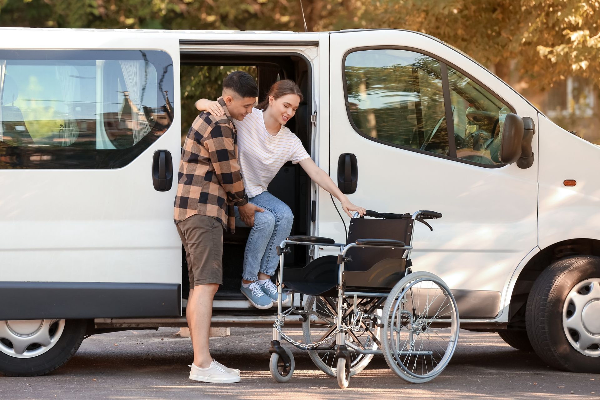 Man helping a person with mobility issues into a white van, wheelchair visible. Outdoors, sunny.