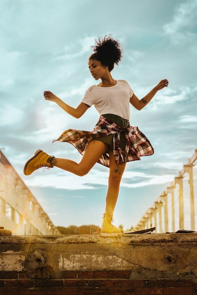 Woman dancing on a rooftop, arms outstretched, wearing a white shirt, plaid skirt, and boots. Blue sky background.