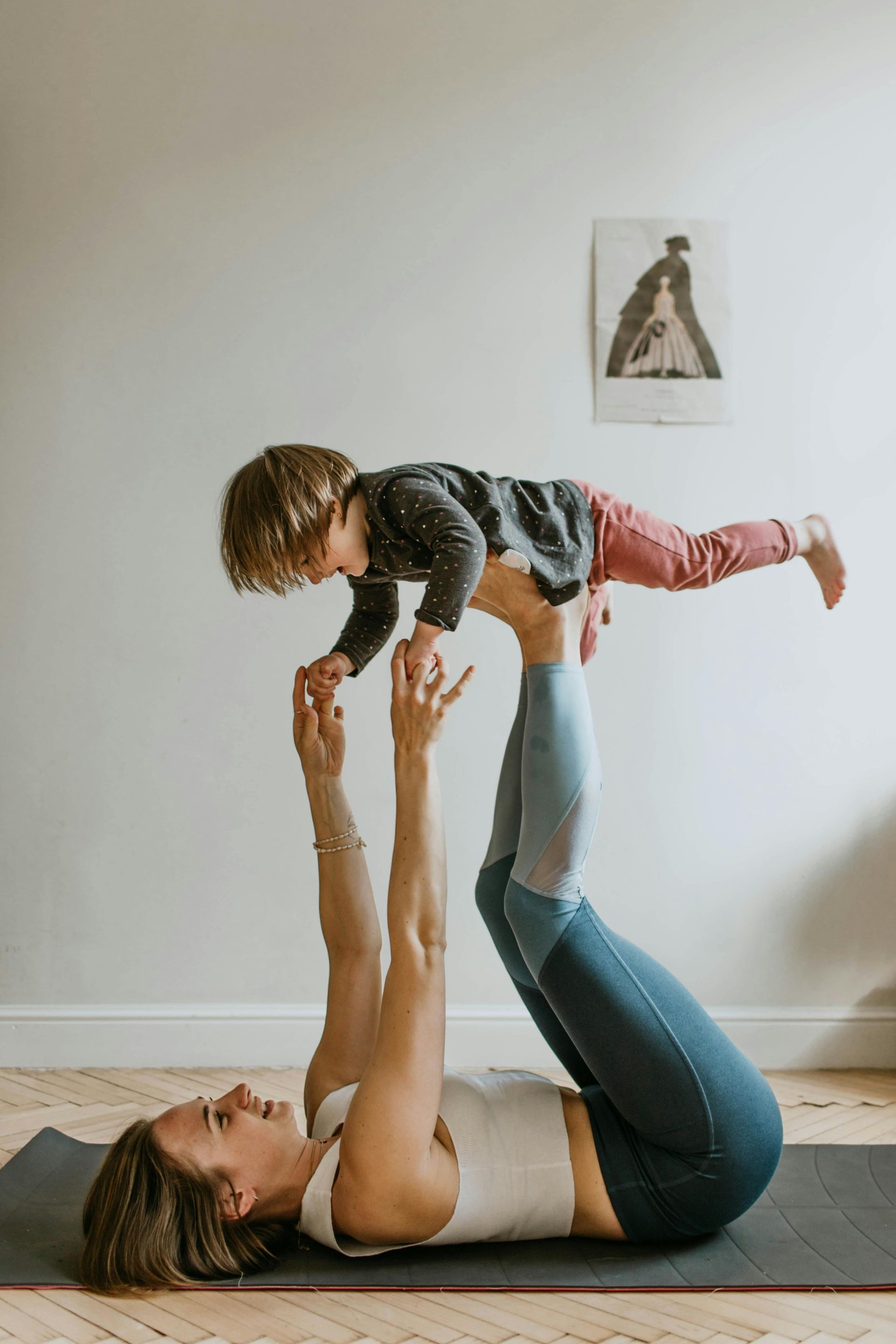 Woman doing yoga, holding up a child in the air. Both are smiling, inside a room.