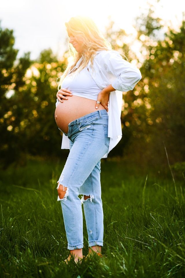 Pregnant person in white top and ripped jeans, touching belly, outdoors in a field with sunlight.