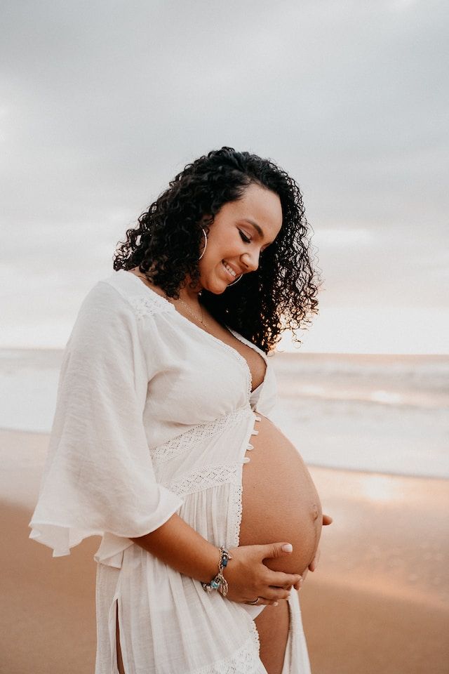 Pregnant person on a beach, wearing a white dress and holding their belly, smiling, with an ocean backdrop.