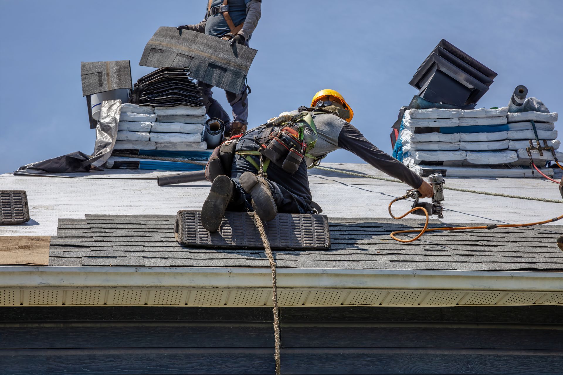 Commercial roofing services crew installing shingles on sloped roof with safety harness and tools.