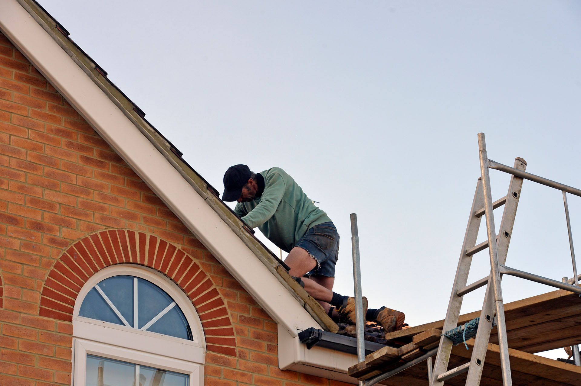 Contractor working on roof shingles of a brick house under clear sky.
