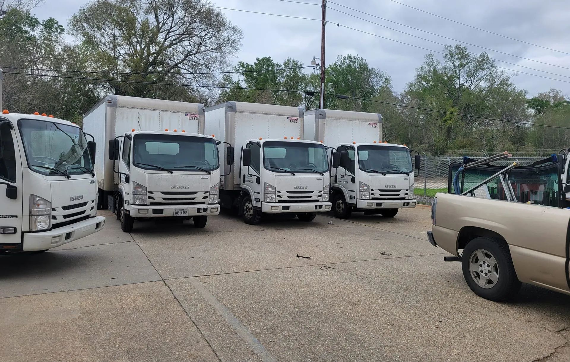 White box trucks parked in a lot, with a pickup truck on the right. Overcast sky.