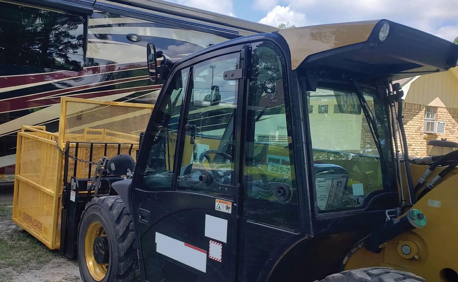 Black and yellow construction telehandler with cage attachment, parked near a recreational vehicle on a sunny day.