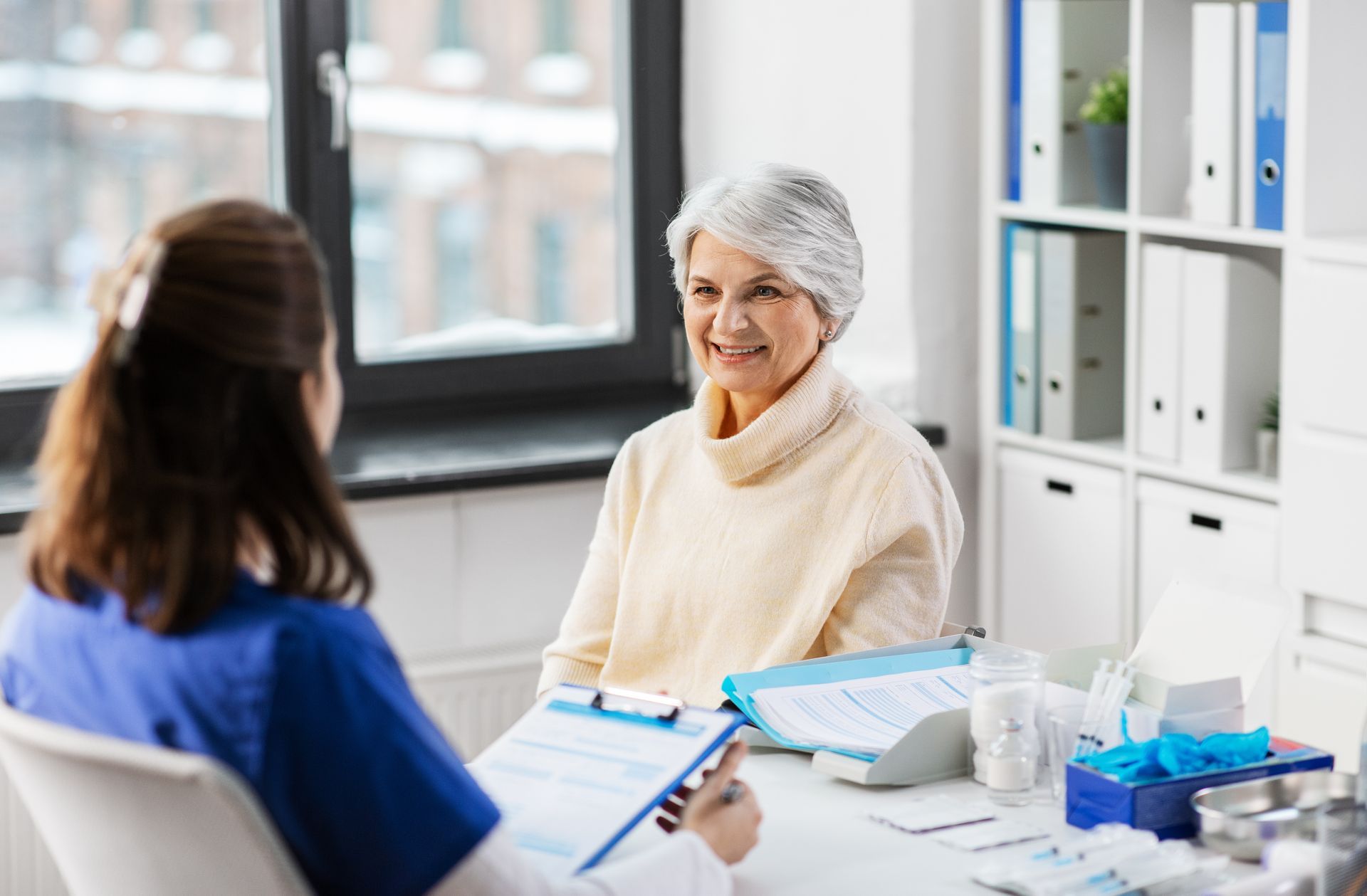 Doctor and patient in an office. Doctor in blue scrubs reviews clipboard, patient smiles. Bright room, window in background.