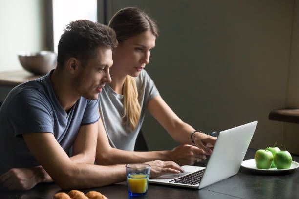 Couple using laptop in a kitchen, looking at the screen, with orange juice and apples on the counter.