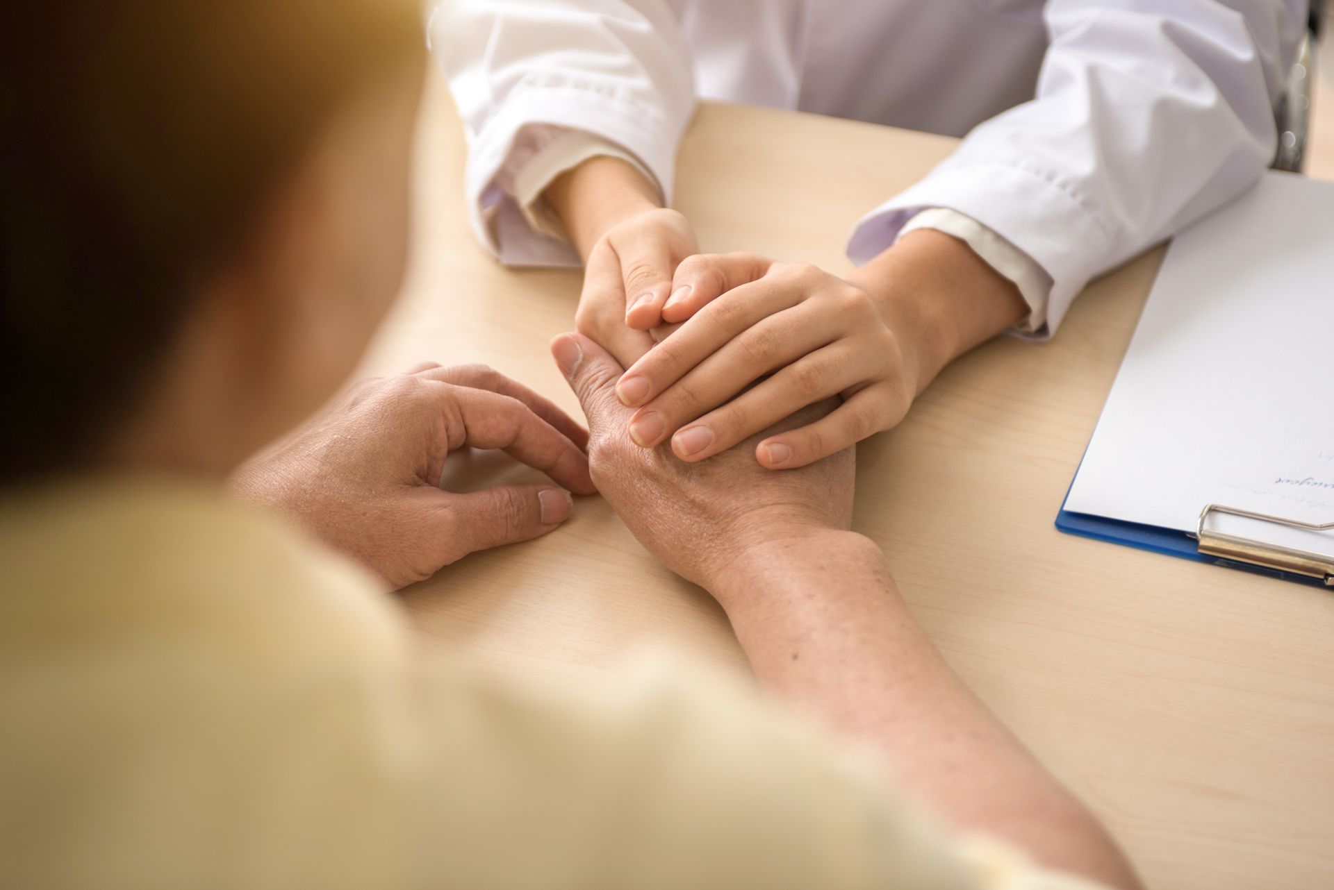 Hands clasped: medical professional comforting a patient at a desk. Clipboard nearby.