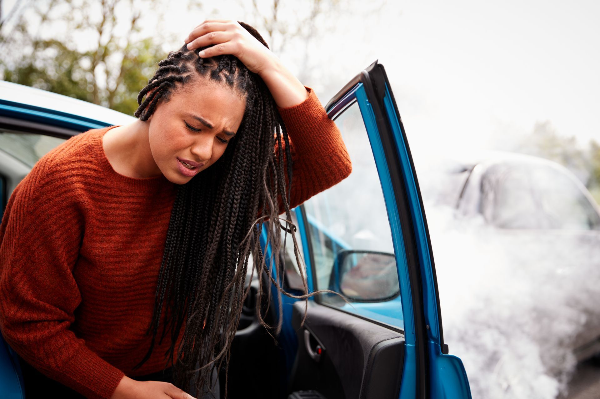 Woman exits a blue car after a collision; smoke rises from a damaged car in the background, appearing distressed.