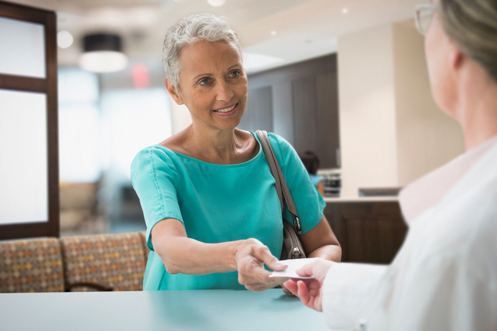 Senior woman giving card to receptionist in modern office lobby with teal shirt and bright decor.