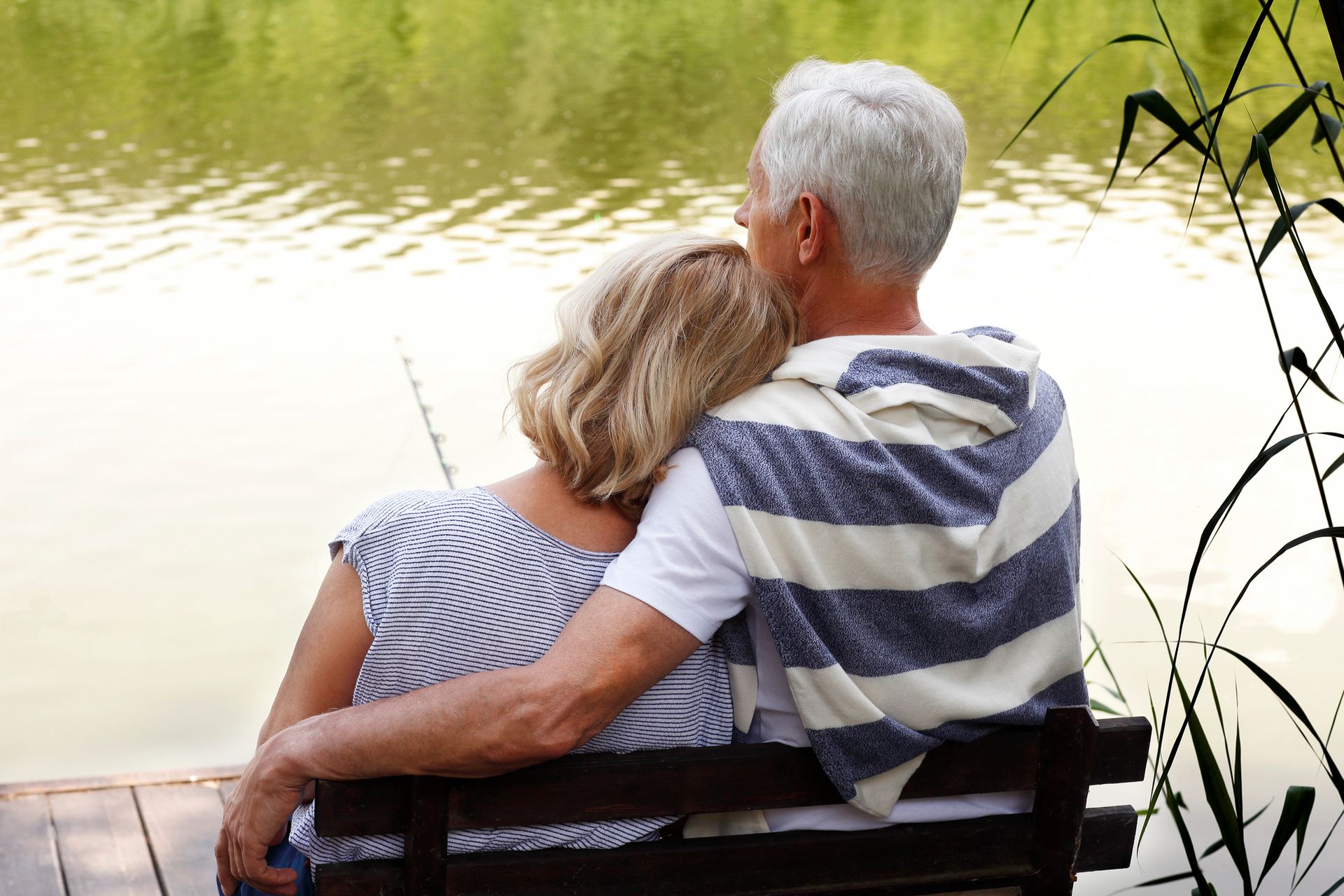 Couple embracing on a bench, looking at a calm lake; partially visible fishing rod.