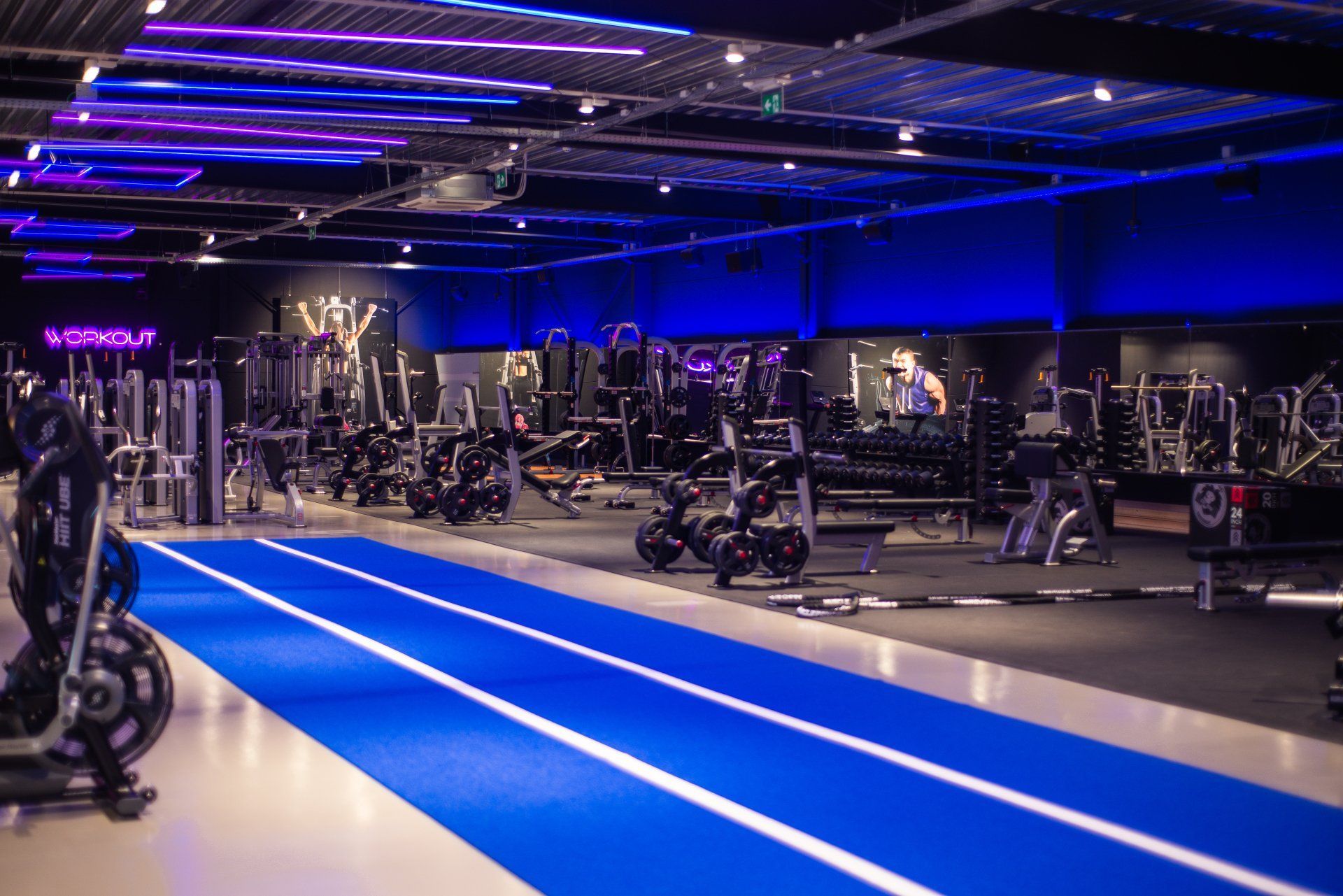 Gym interior with a blue running track and exercise equipment. Dark walls with neon lights.