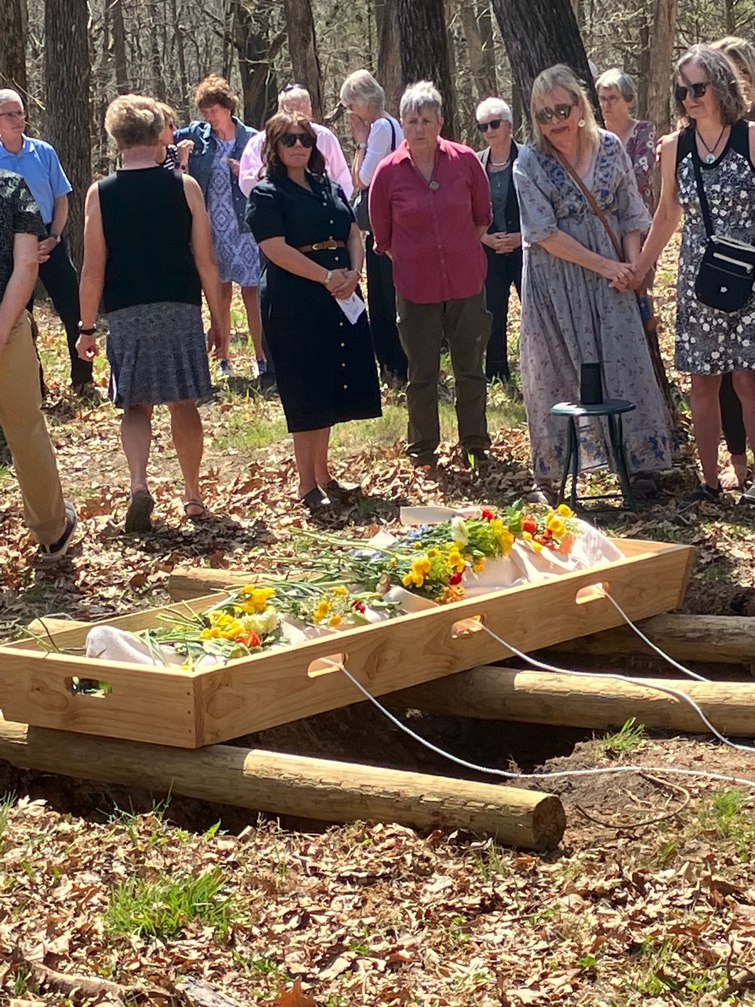 A group of people are standing around a wooden coffin filled with flowers.