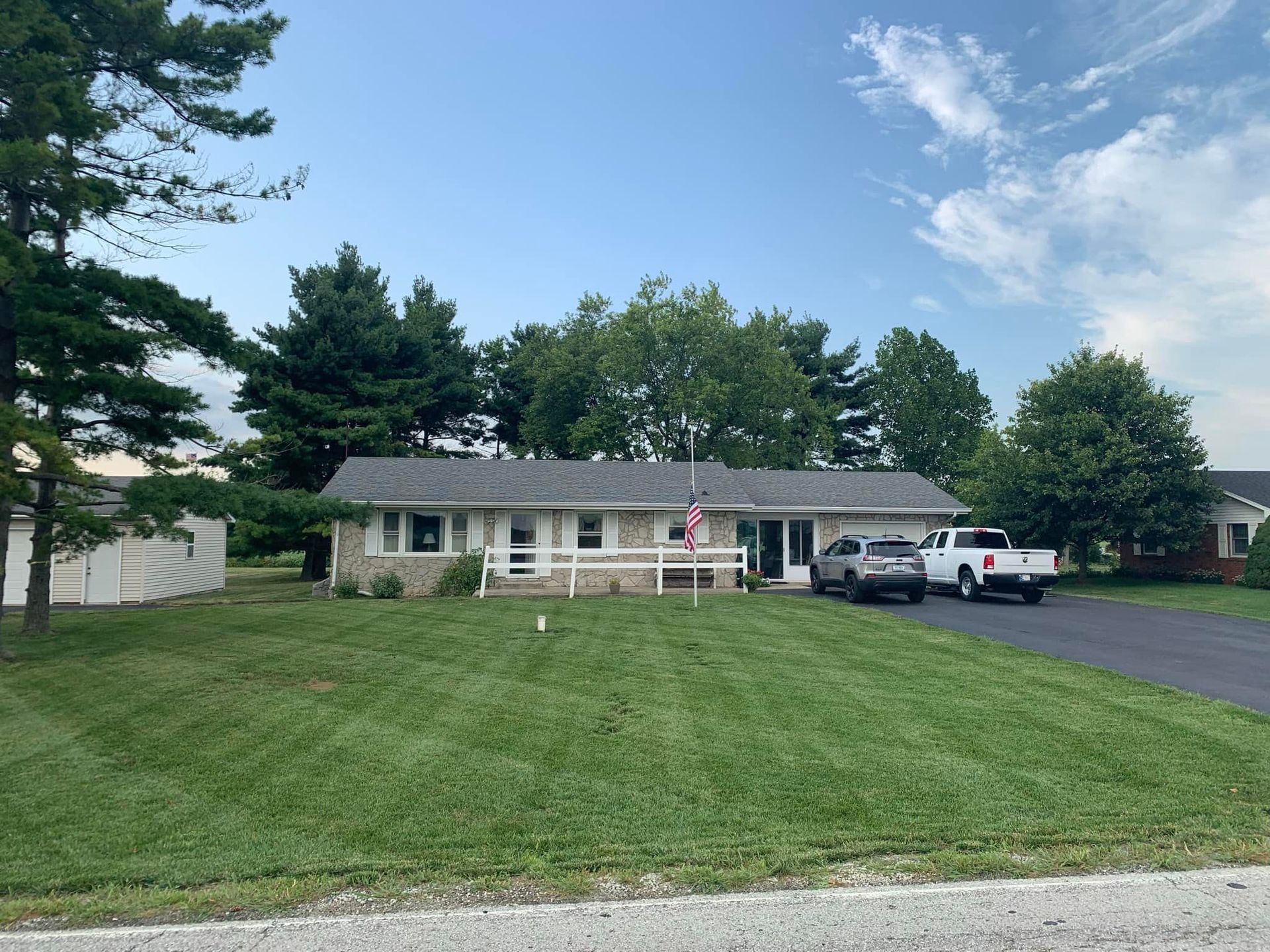 Ranch-style house with stone facade, American flag, vehicles in driveway, green lawn, trees, blue sky.