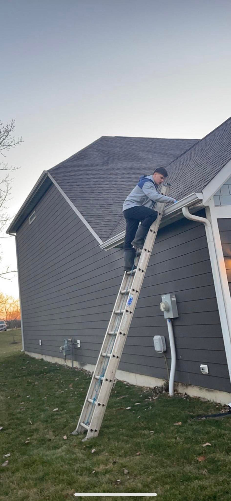 A person on a ladder cleaning a house gutter, a gray-sided house, and a grassy yard.