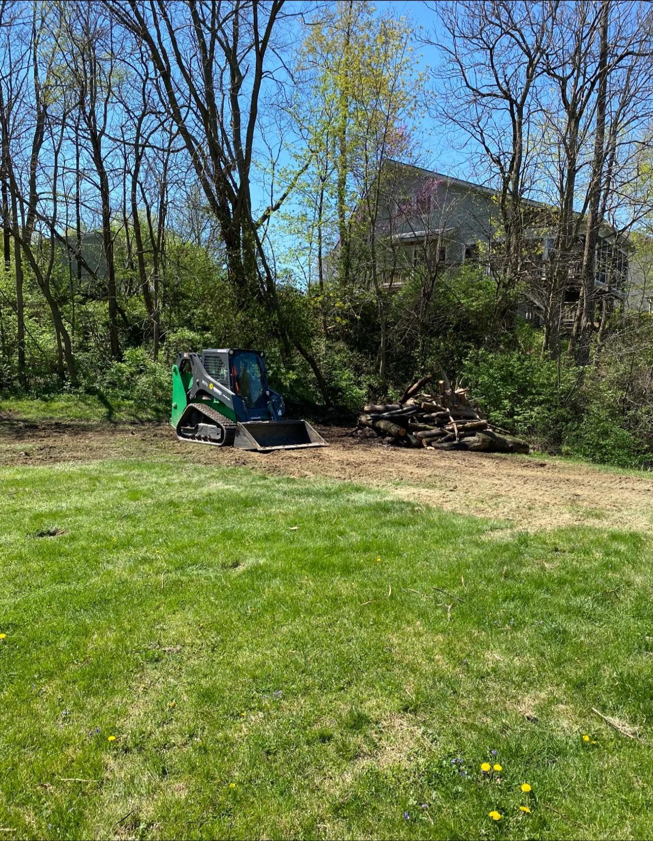 Green and black skid steer on dirt clearing, logs, trees, house in background. Sunny day.