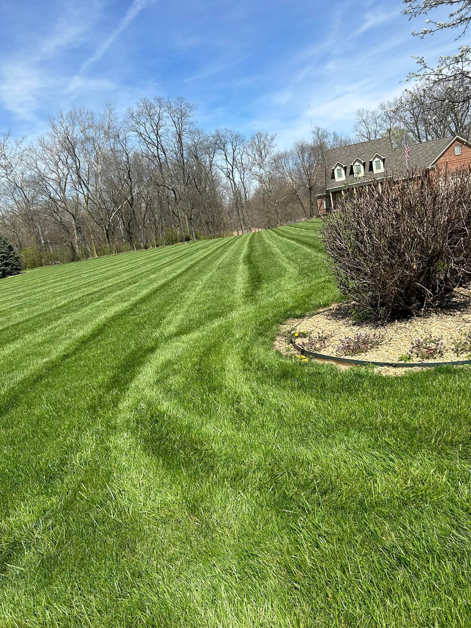 Green lawn with mowing stripes on a hillside, with trees in the background and a house on the right.
