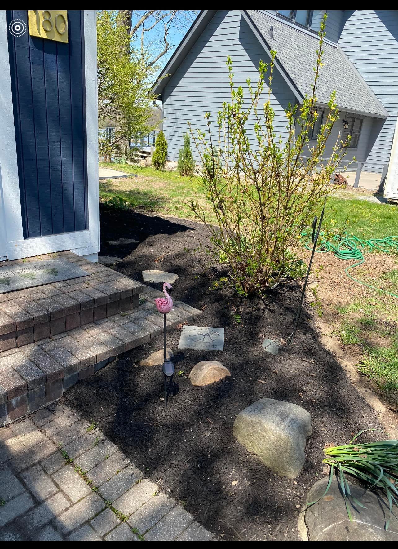 A house with a brick walkway and mulch garden, featuring stepping stones and a pink flamingo.