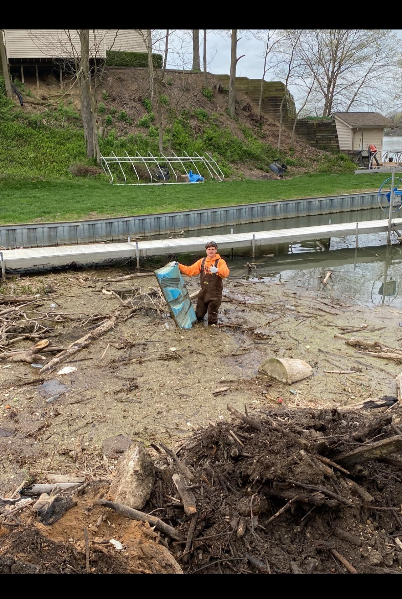 Man in waders holds a blue item while standing in a muddy, drained lakebed by a dock.