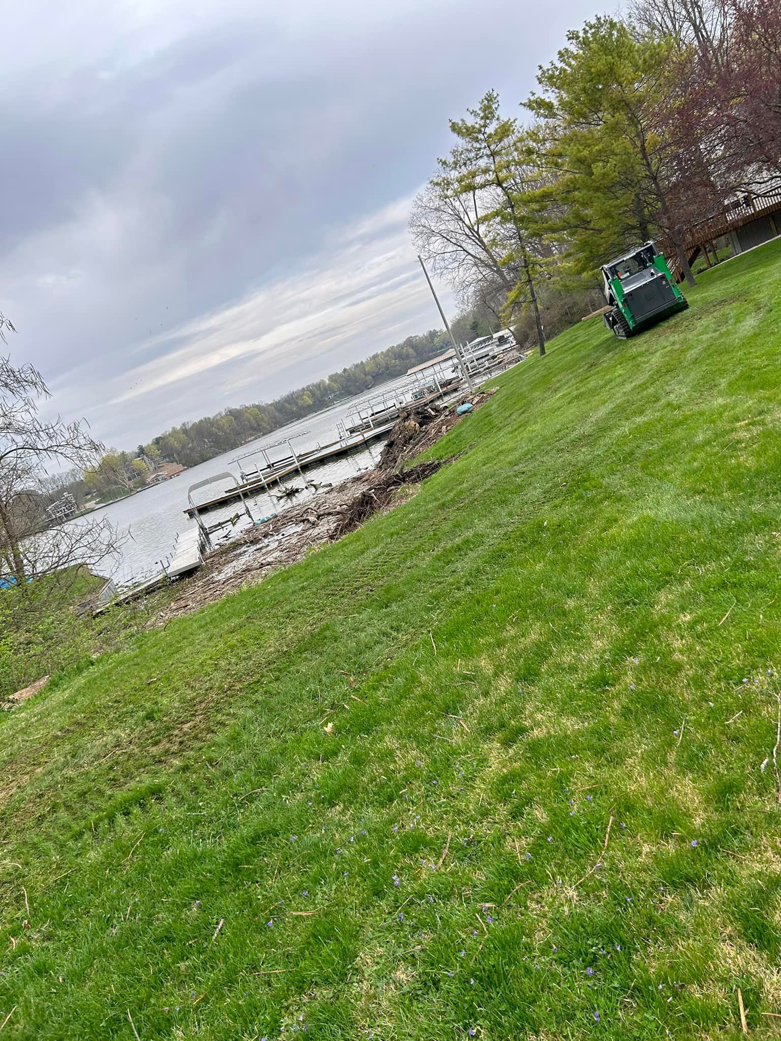 Green lawn slopes down to a lake, with a green machine and trees visible. Cloudy sky.