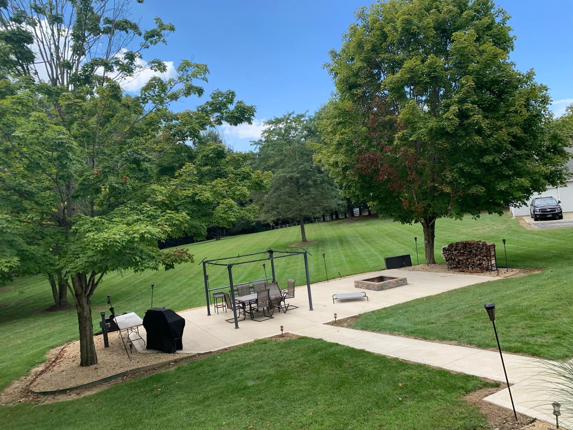 Backyard patio with grill, seating, fire pit, and stacked firewood. Lush green lawn, trees, and blue sky.