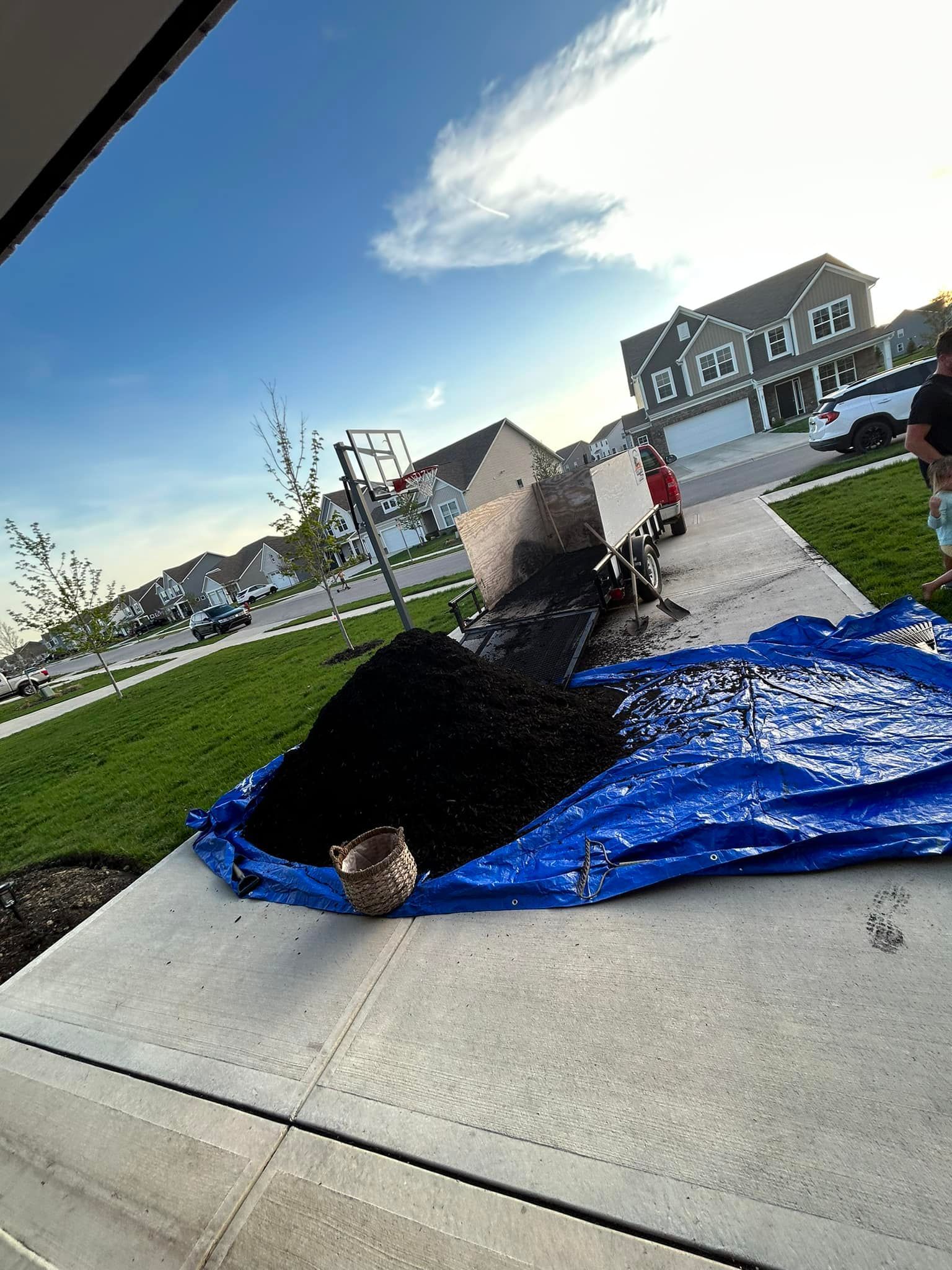 Black mulch spilled on a blue tarp on a sidewalk; person standing in the background. Houses and grass visible.