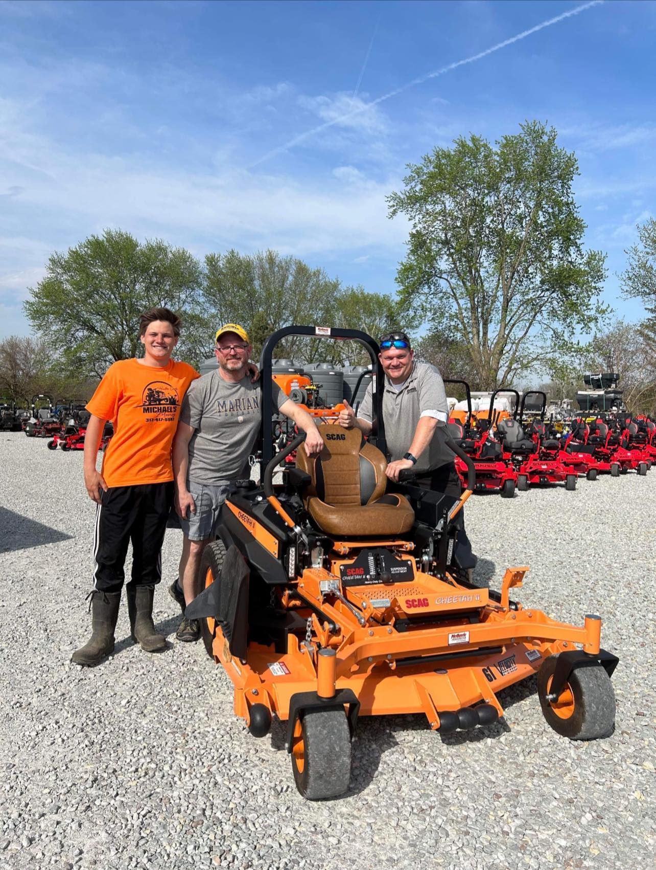 Three people pose with an orange lawnmower. Outdoors, sunny. One person sits on the mower. Other lawn equipment visible.