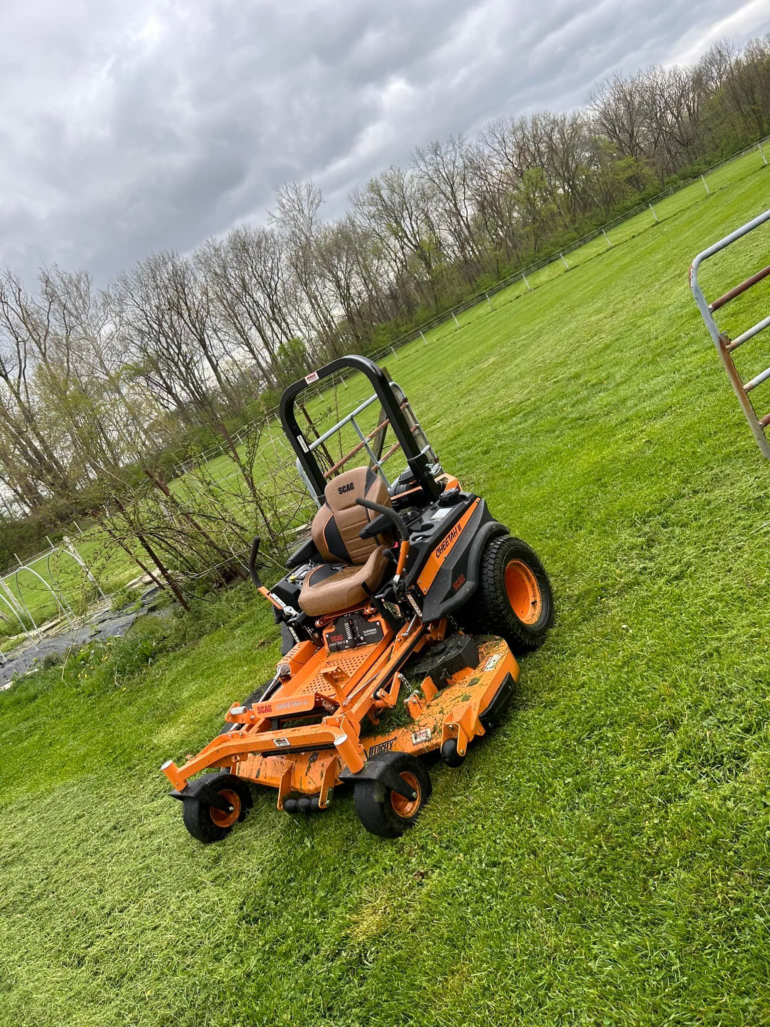 Orange and black zero-turn mower on green grass. Overcast sky in background.