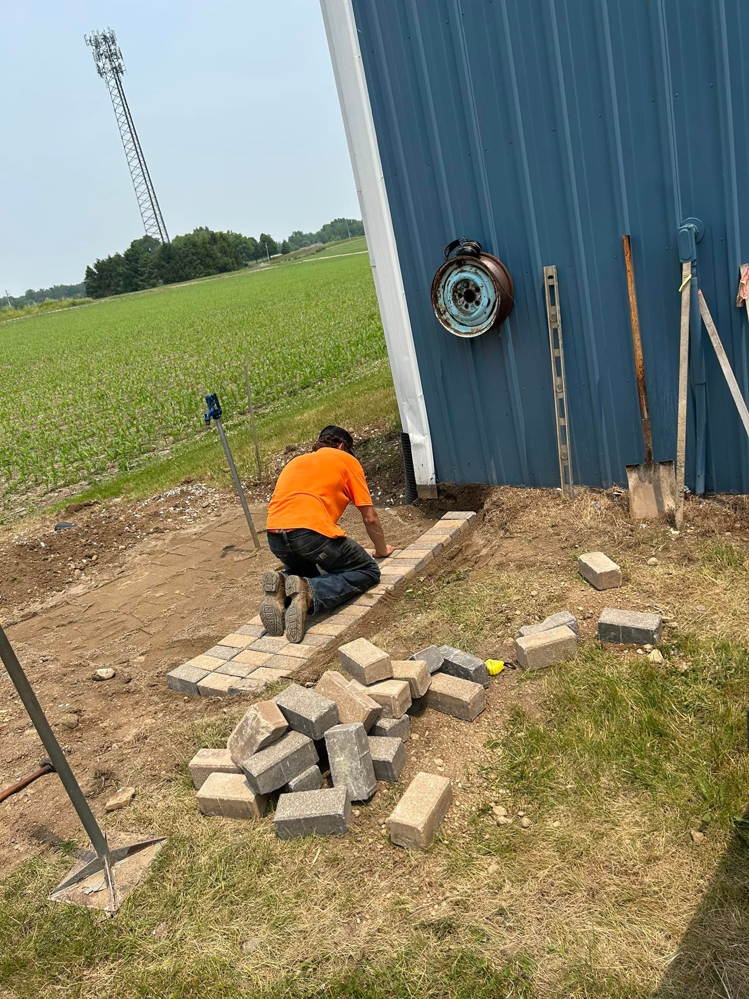 Person in orange shirt laying bricks near blue building in field.